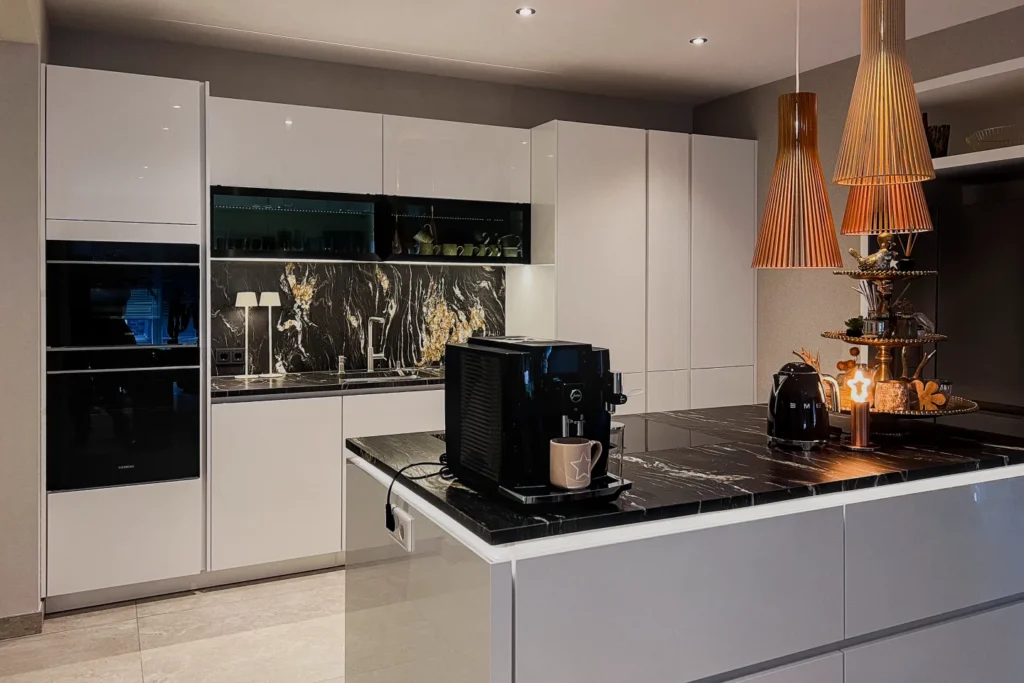 Moderne Küchenzeile mit weißen Schränken, Kochinsel, Kaffeemaschine und Pendelleuchten sowie dunkler Fliesenrückwand