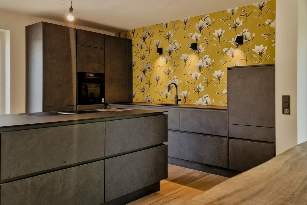 Moderne Küchenzeile mit dunklen Fronten, gelber Blumen-Rückwand, Spüle und eingebautem Backofen, Holzfußboden.