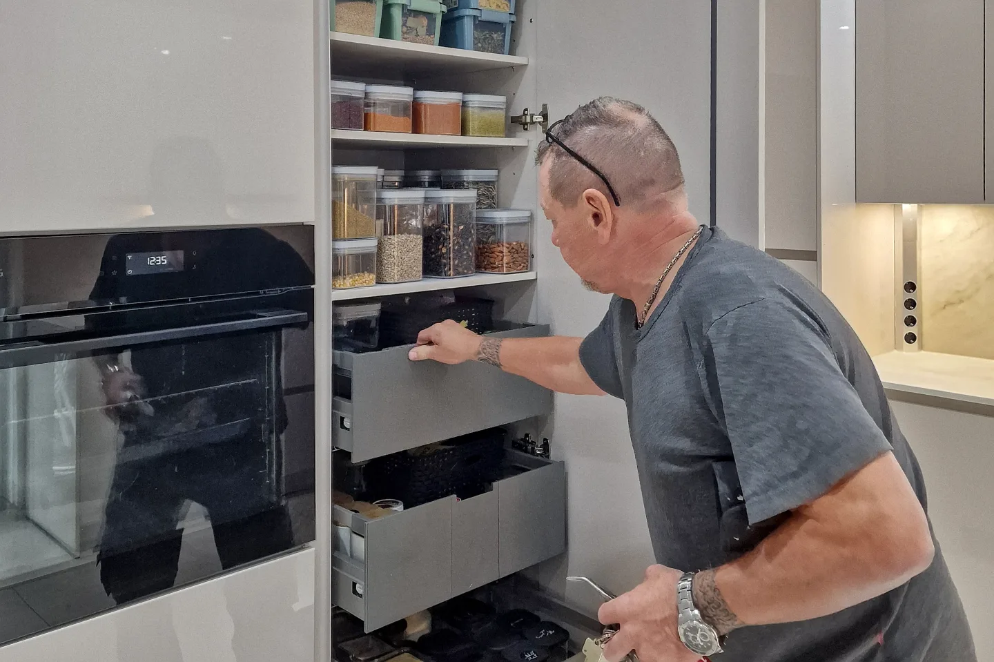 Mann richtet einen Küchenschrank mit herausziehbaren Vorratsfächern ein, neben eingebautem Ofen.
