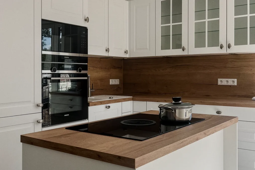 Weiße Einbauküche mit Backofen, Kochfeld und Topf auf Holzarbeitsplatte vor Holzpaneel an der Wand.