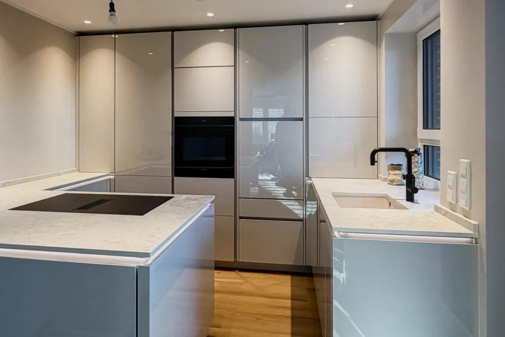 Moderne Einbauküche mit Hochschränken, Backofen, Kochfeld und Spüle an einer Kücheninsel.