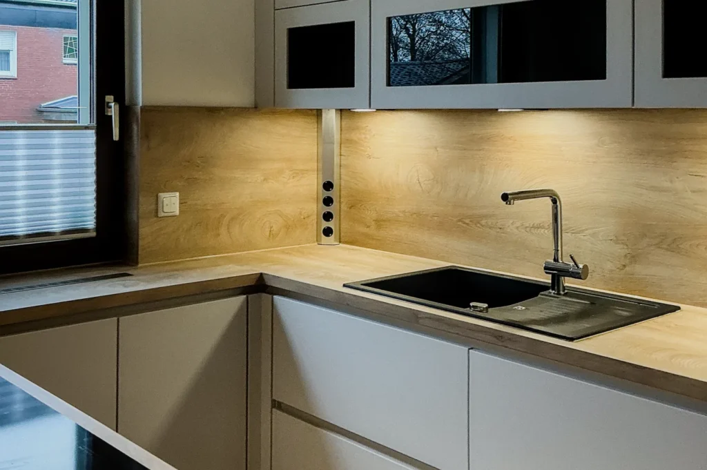 Moderne Küchenzeile mit Spüle, Armatur und Arbeitsplatte, Holzrückwand sowie Schränken und Einbausockel.