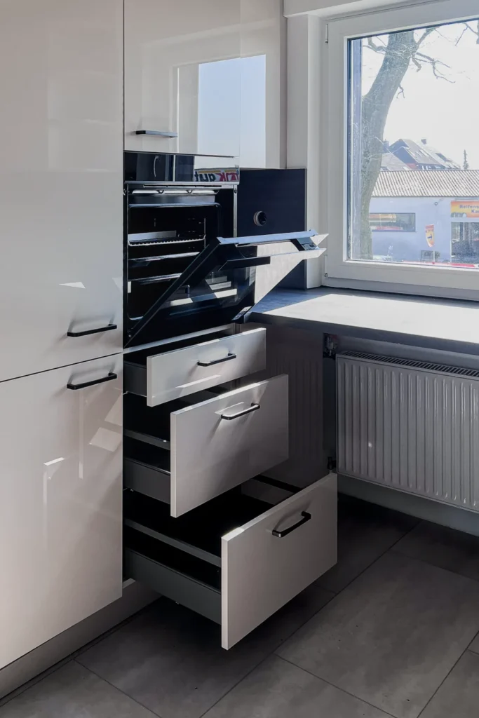 Küchenzeile mit Backofen, Schubladen und Herd neben dem Fenster in einer hellen Einbauküche.