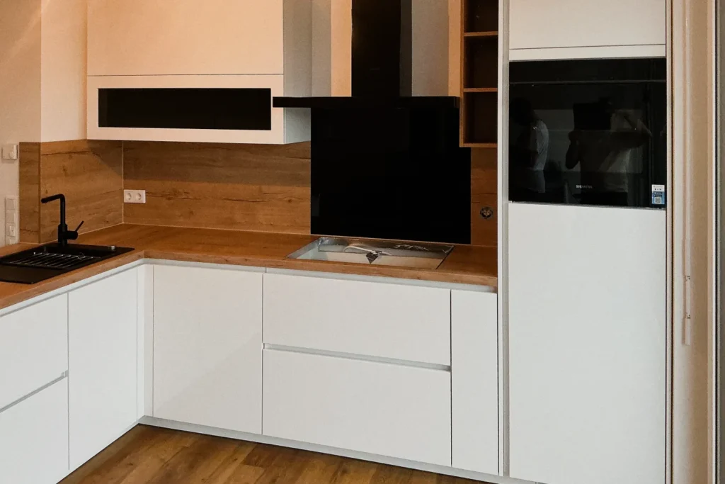 Weiße Küchenzeile mit Holzarbeitsplatte, Herd, Spüle und integrierter Spülmaschine, daneben Backofen mit Glastür.