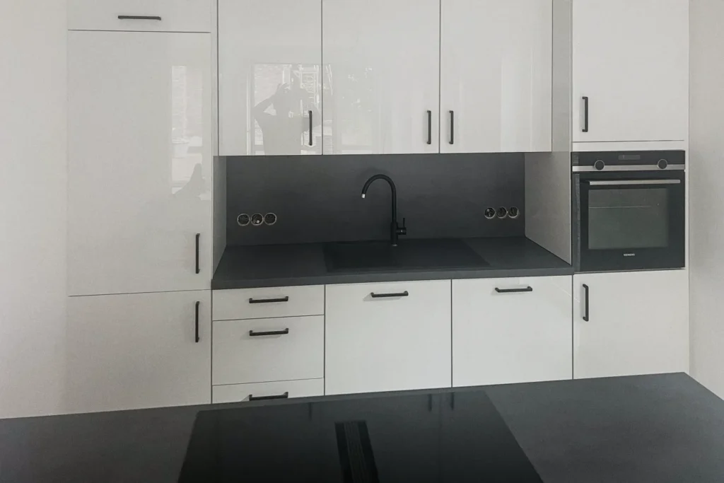 Weiße Küchenzeile mit dunkler Arbeitsplatte, Spüle mit Armatur und eingebautem Backofen, Schränke mit Griffen.