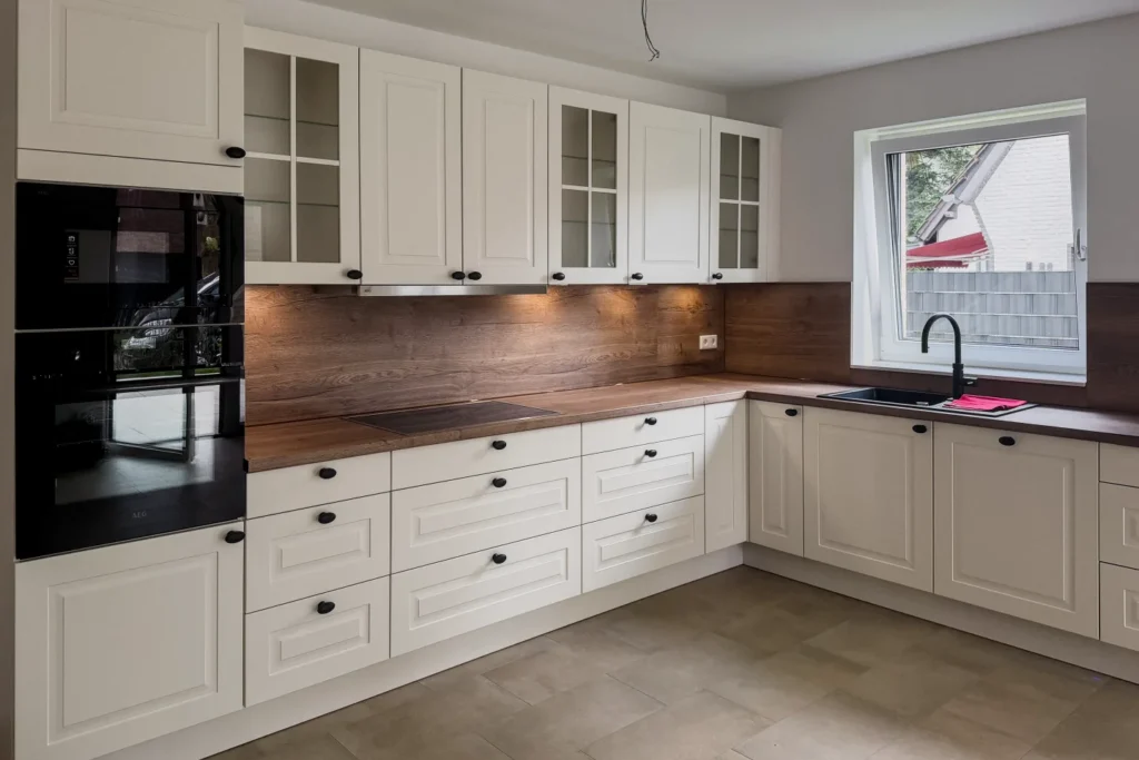 KU_017: Helle L-förmige Küchenzeile mit Hochschränken, Glastüren, Backofen und Spüle am Fenster, Holz- und Fliesenboden.