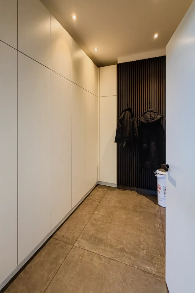 Flur mit weißem Einbau-Schrank, dunkler Garderobenwand mit Haken, Spotlights und Fliesenboden.