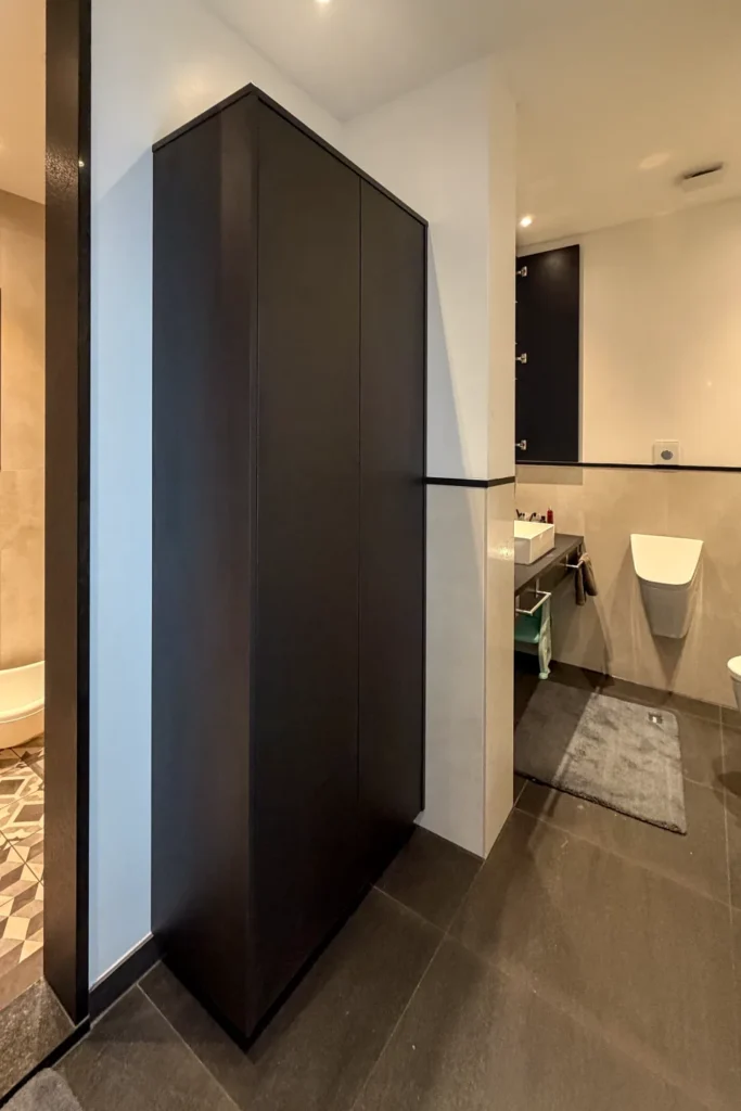Schwarzer Hochschrank in einer modernen Badezimmernische mit Waschbecken, Handtuchhalter und Toilette im Hintergrund.