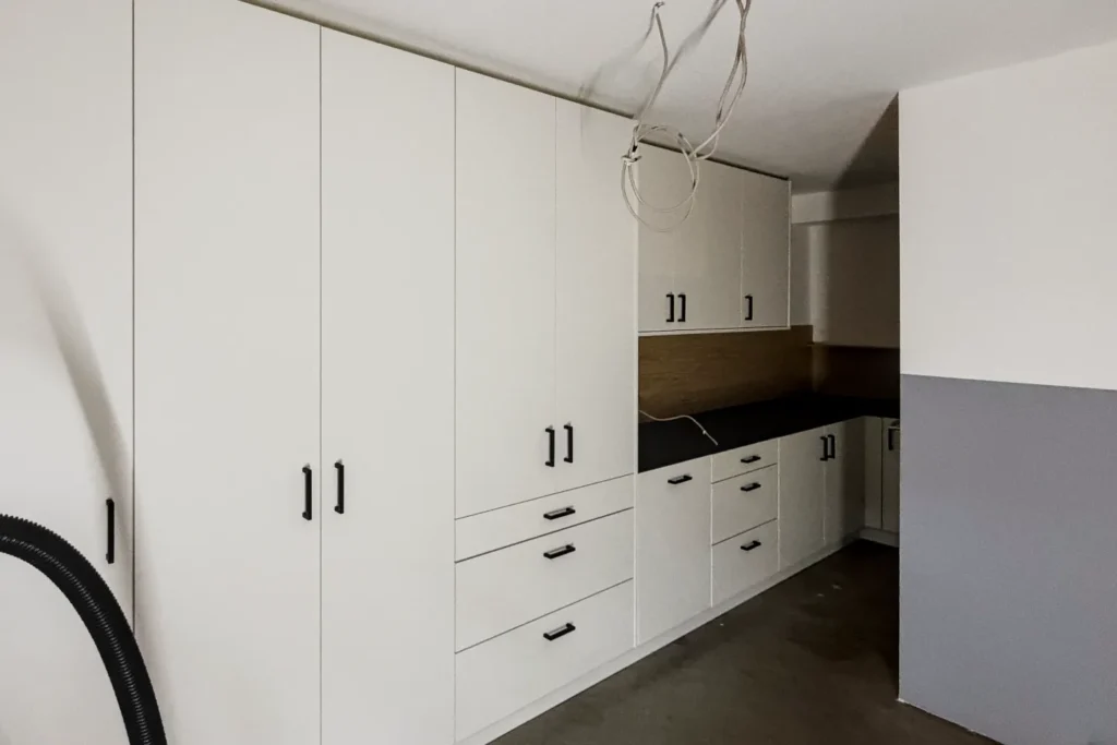 Weiße Einbauküche mit Hochschrank, Hängeschränken und Schubladen; daneben offene Nische und Elektroanschlusskabel.