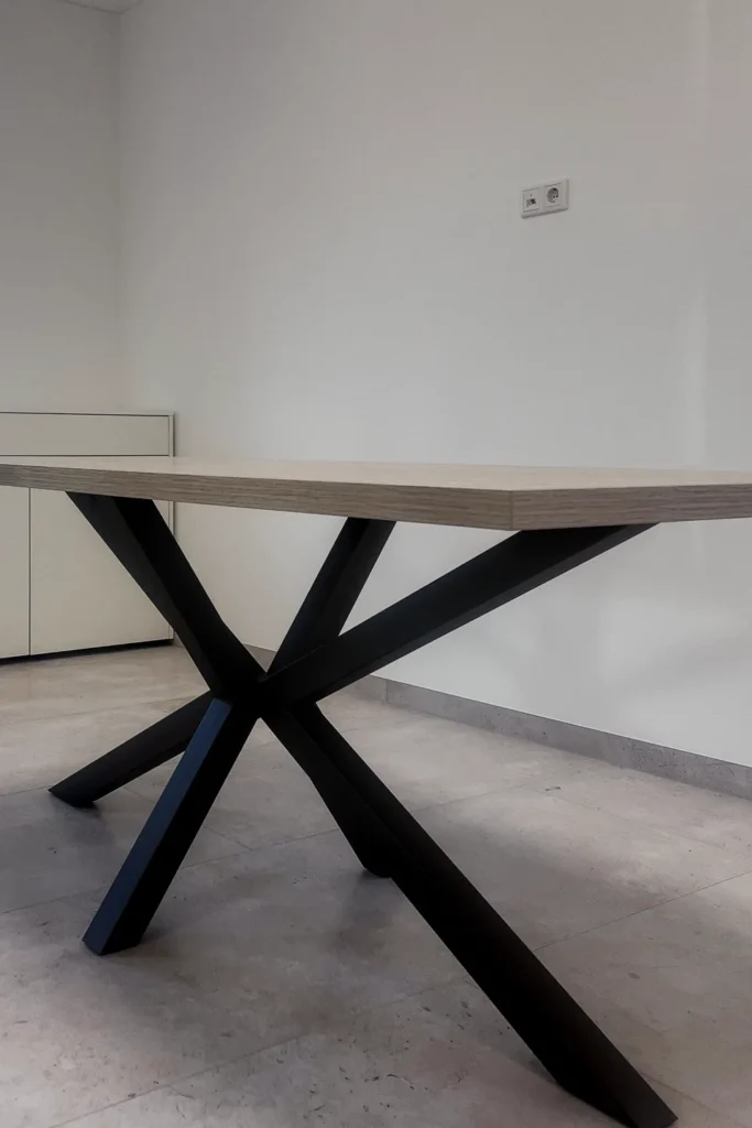 Esstisch mit dunklem X-förmigem Metallgestell und beiger Platte in heller Küche, Wandsteckdose sichtbar.
