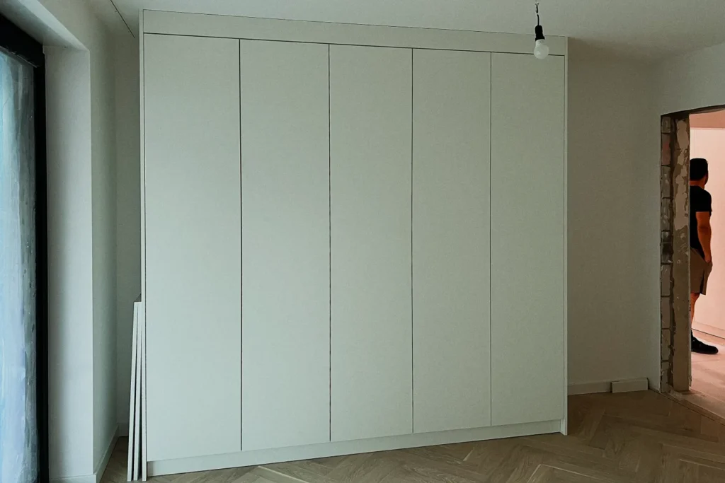 Weiße Schrankwand mit mehreren Türen an der Wand, Holzfliesenboden und Deckenlampe im Hintergrund.