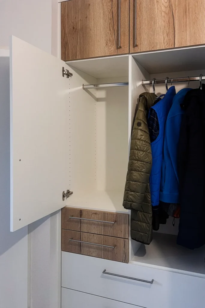 Einbauschrank mit Holzfronten und Schubladen; im Innenraum hängen Jacken an Garderobenstangen.