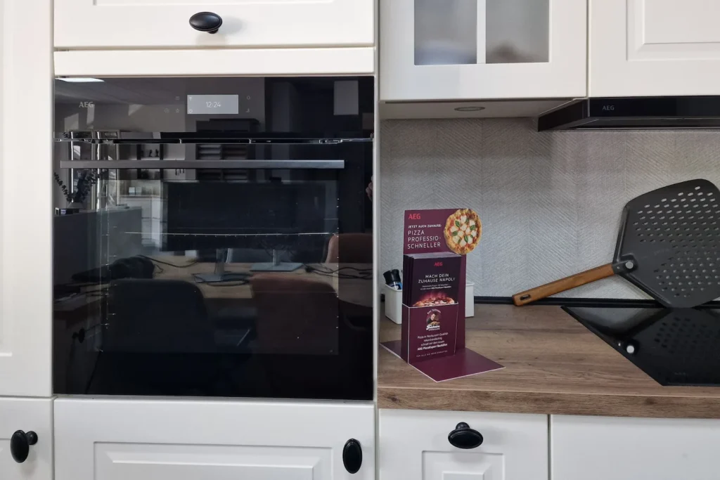 Einbaubackofen mit geöffneter Tür, daneben Küchenarbeitsplatte mit Pizzaback-Box „AEG“.
